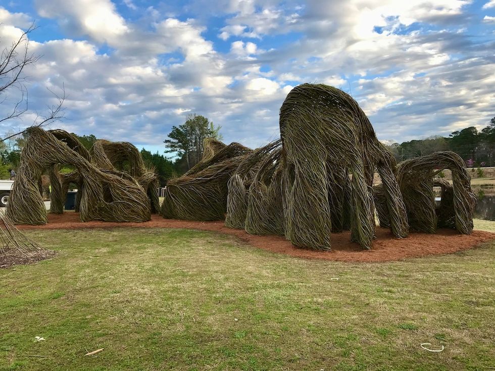 Woven stick sculpture on grass under a cloudy sky, surrounded by trees.