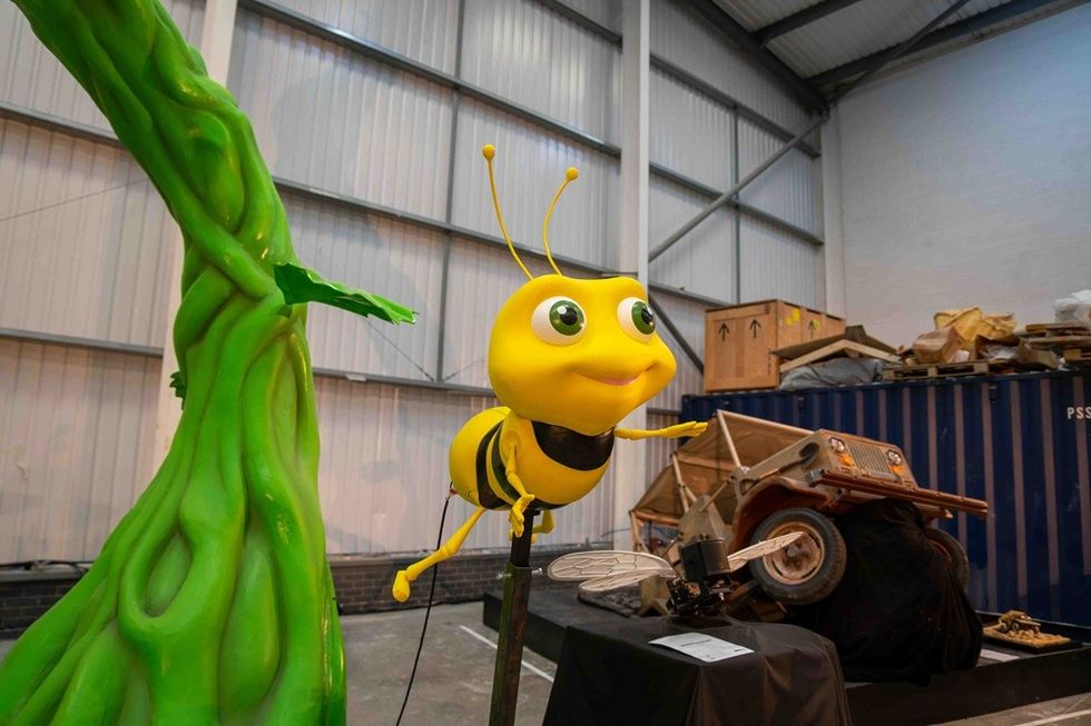 Yellow cartoon bee statue beside green plant in a storage area with a vintage car.