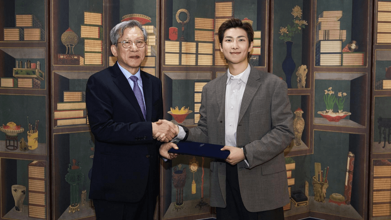 You Hong June and RM shaking hands in front of a colorful bookshelf backdrop.