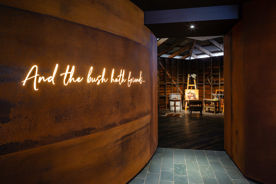 Rust-colored wall leading into gallery with lit text "and the bush hath friends"