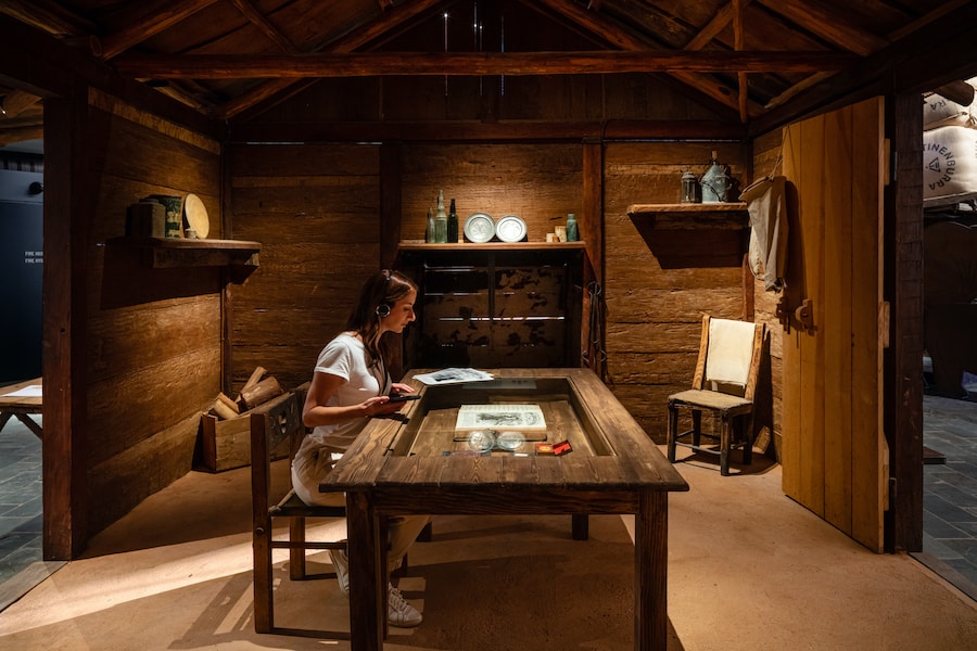 A woman sits at an interactive table inside a cabin exhibit