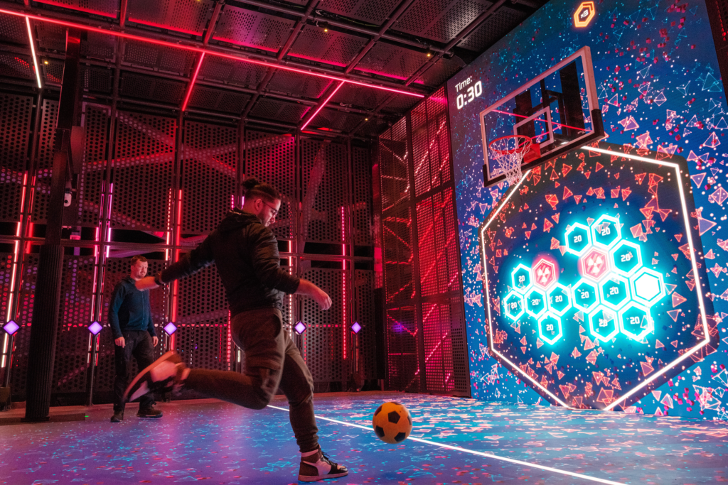 Two men playing Wall Ball on Glow Court, an interactive LED sports court aboard Norwegian Aqua. One player is mid-kick, aiming a yellow and black football towards a large digital screen displaying hexagonal targets, points, and a basketball hoop. The futuristic court is lit with vibrant neon red and blue lights, creating an immersive, tech-enhanced gaming experience on the ship’s top deck at sea.