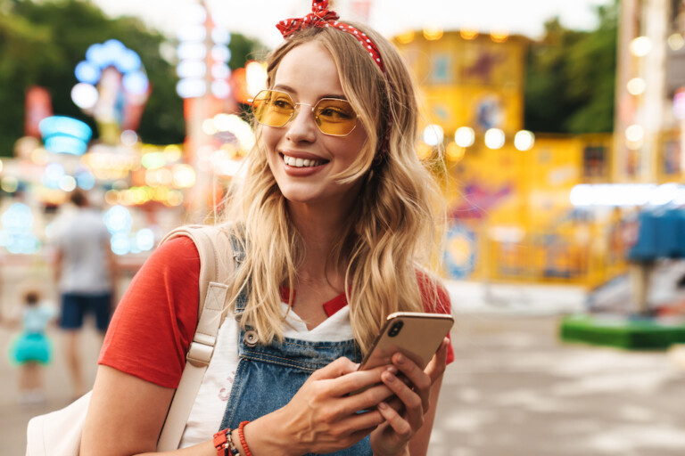 Guest data Image of a smiling young cheery blonde woman in amusement park using mobile phone.